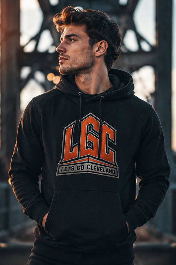 A man wearing a black pullover hoodie with a large orange and white 'LGC LETS GO CLEVELAND' graphic on the chest, standing in front of a blurred bridge structure at sunset.